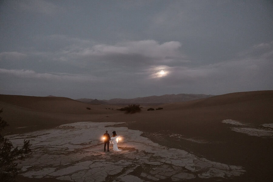 Wide shot of the couple holding lanterns in the dark desert with cracked ground and a glowing moon.