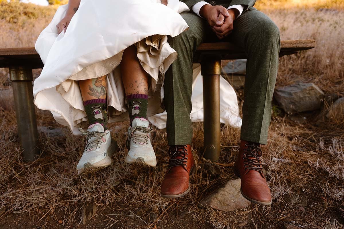 A couple sitting on a bench with the woman wearing hiking shoes and fun socks under her wedding dress, while the man wears dress shoes.