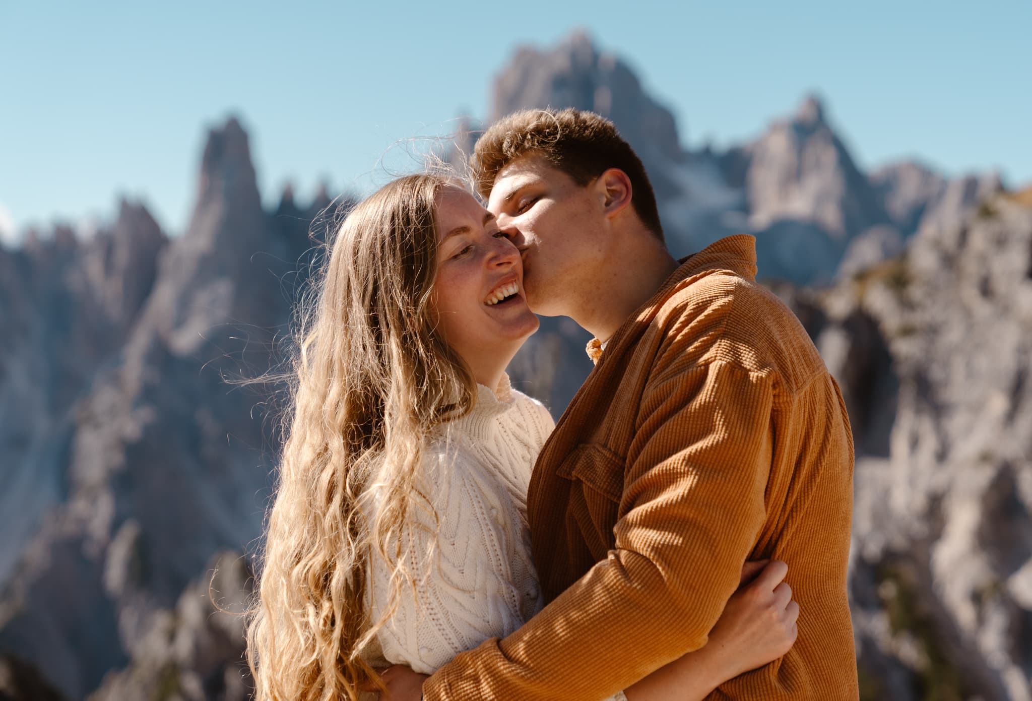 Boyfriend kissing girlfriend on the cheek with mountains in the background. Girlfriend is laughing.