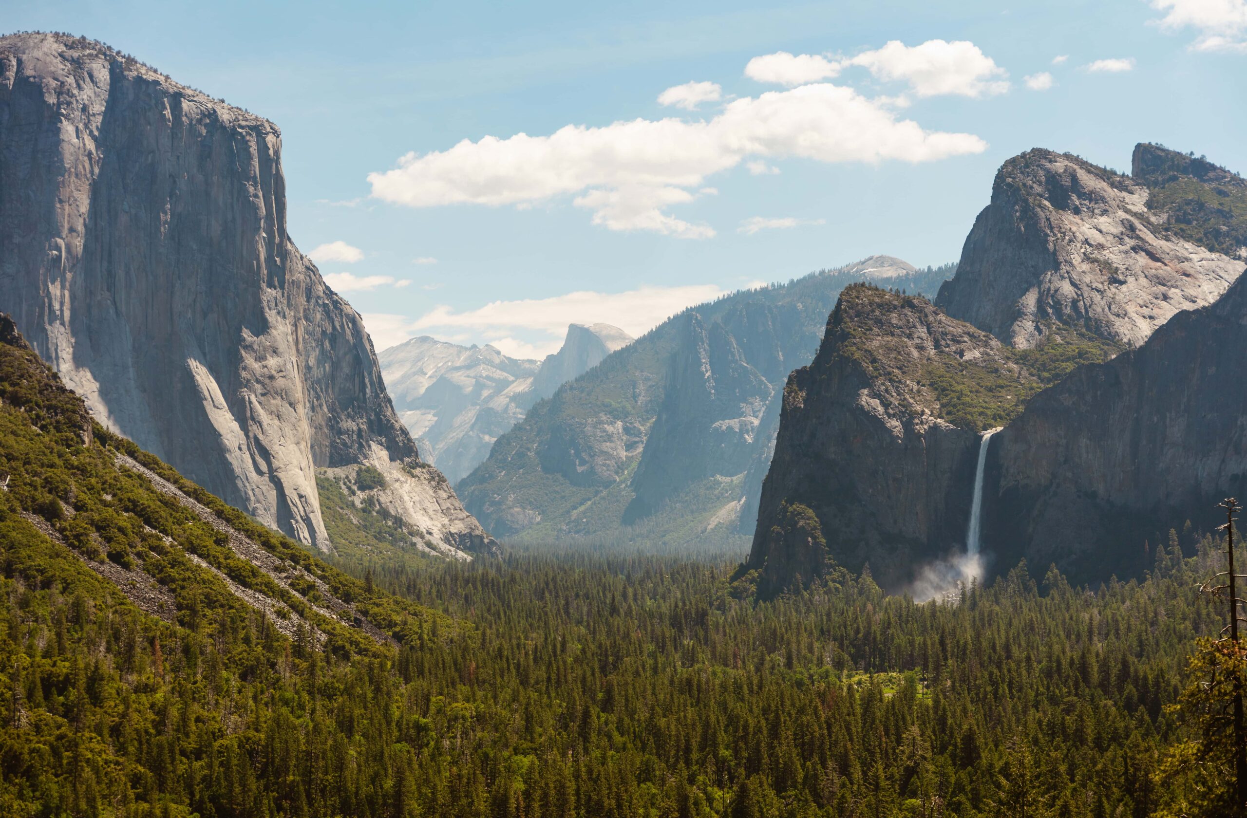The image shows a scenic view of Yosemite Valley, with El Capitan on the left, Bridalveil Fall in the center, and the Half Dome in the distant background, under a blue sky with scattered clouds.