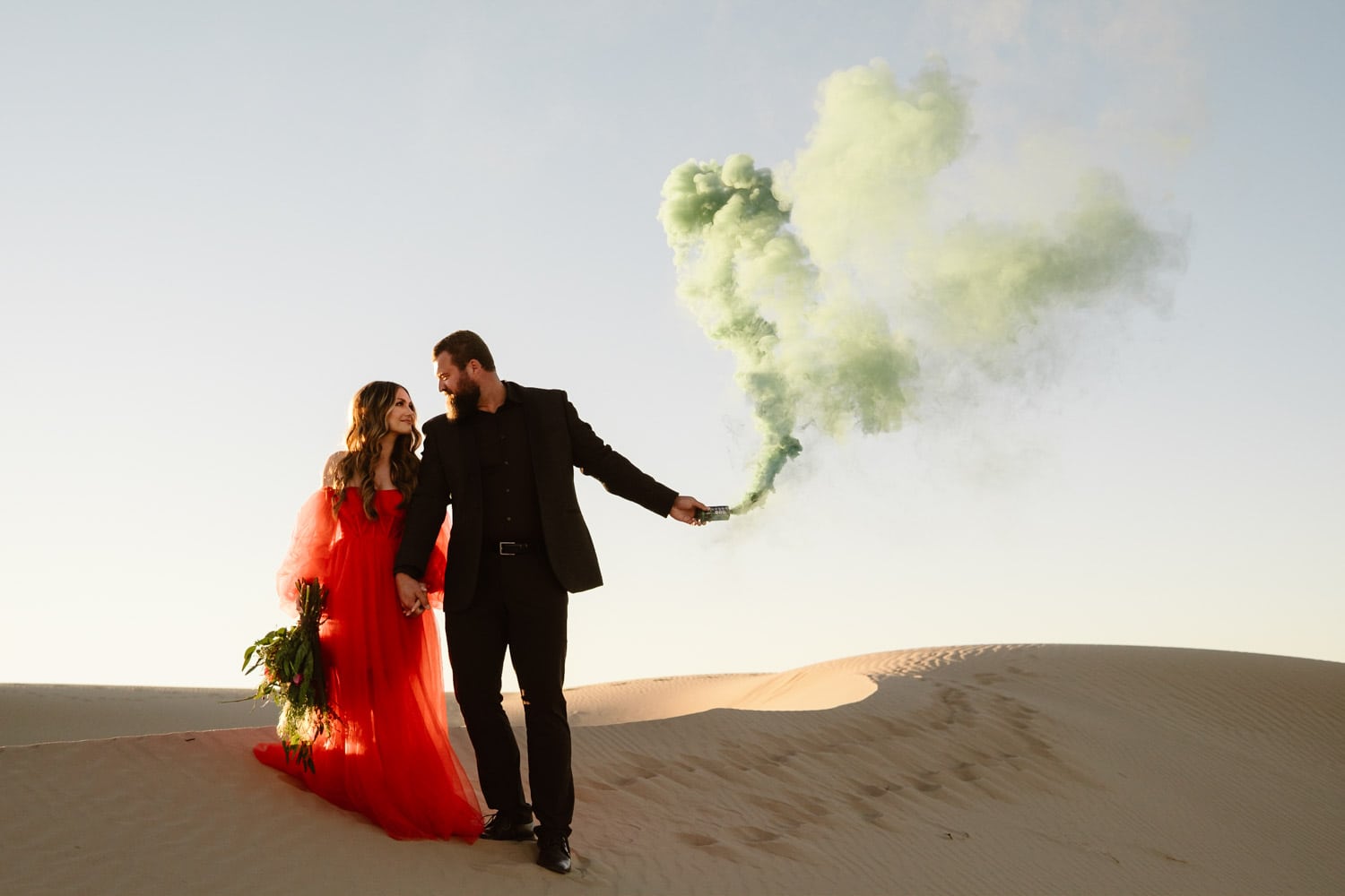 A couple stands hand in hand on the Glamis Sand Dunes with a billowing green smoke flare behind them, the woman in a vibrant red dress and the man in a dark suit.