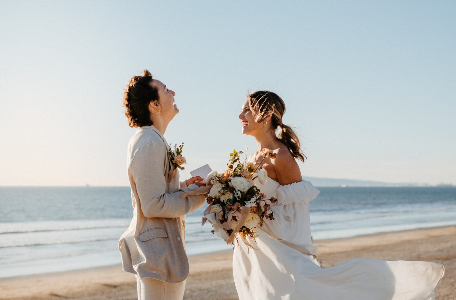 Two brides are laughing together on a beach, with the bride holding a bouquet and her dress blowing in the wind.