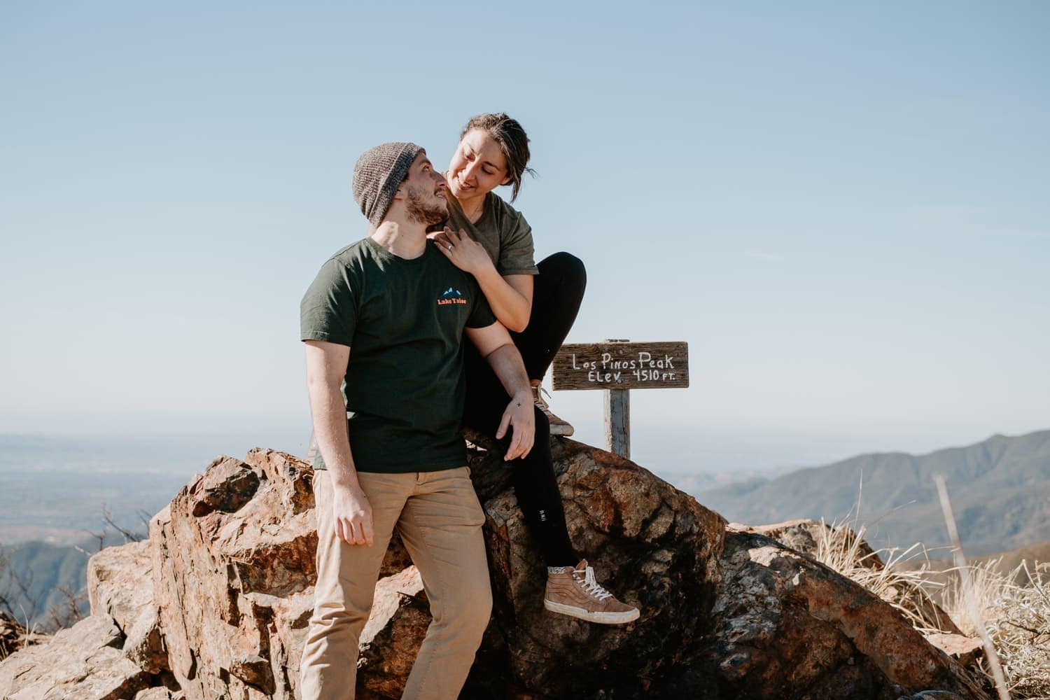 A couple is pictured at Los Pinos Peak, which is at an elevation of 4510 feet, as indicated by the sign. They are enjoying a sunny day outdoors, with the man standing and the woman seated on a rock, her legs draped over his. They appear to be sharing a light-hearted, affectionate moment against a backdrop of distant mountains and clear skies.