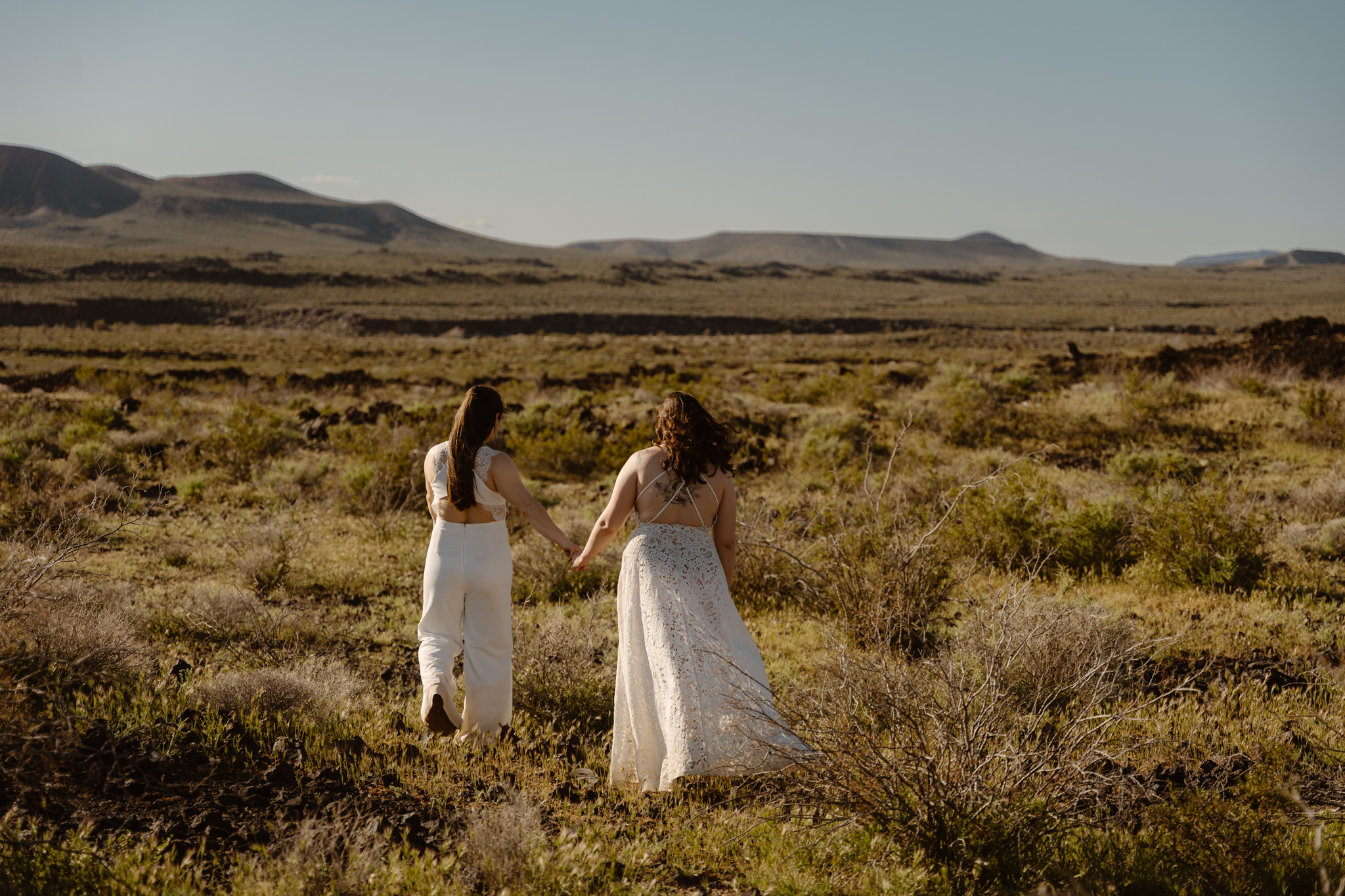 Two people holding hands and walking through a desert landscape, dressed in wedding attire.