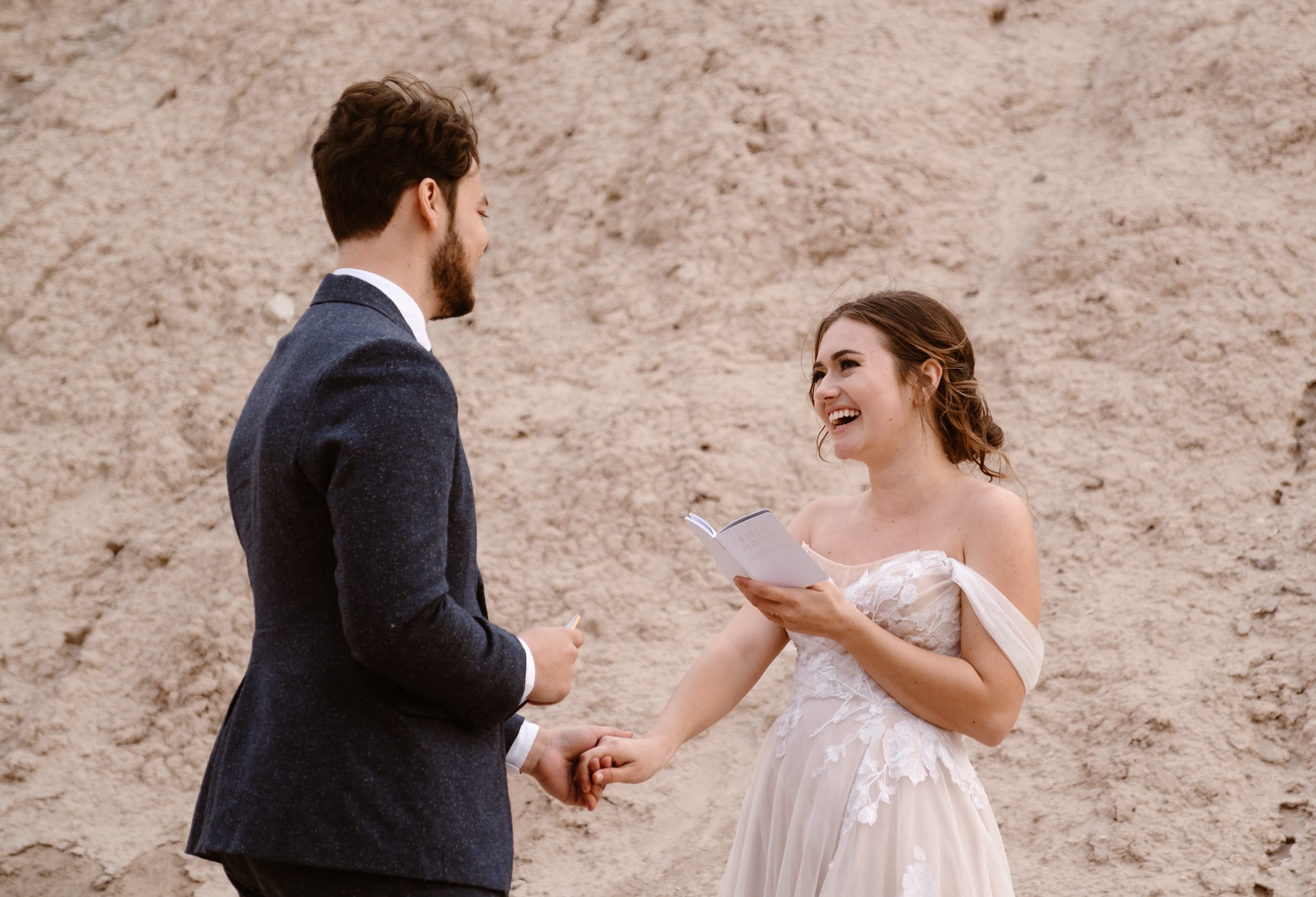 A person in a suit faces a smiling bride holding a vow book, both standing in front of a textured sandy hill.