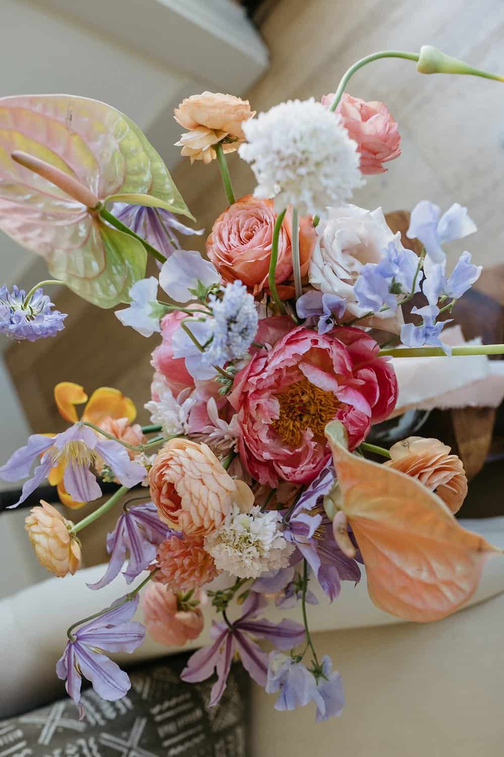 A colorful floral arrangement featuring a mix of pink peonies, orange roses, white blooms, and purple flowers, with some leaves showcasing a hint of pink, is displayed indoors.