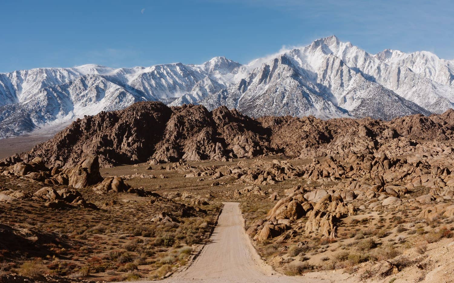 A dirt Movie Road meanders through Alabama Hills, leading towards the snow-covered peaks of a mountain range under a clear blue sky with a crescent moon visible above.