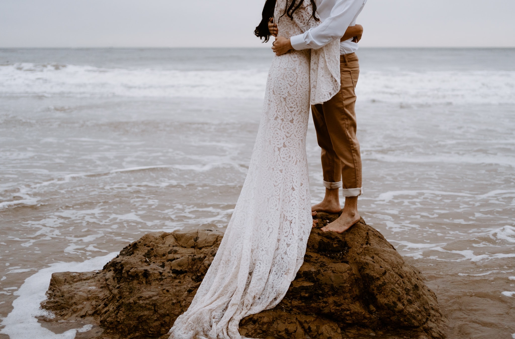 A detailed view of a couple standing on a rock, surrounded by the sea, focused on their embrace and the woman's lace wedding dress.