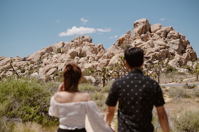 A couple holding hands and walking through Joshua Tree's Hidden Valley, with Joshua trees and mountains in the background.