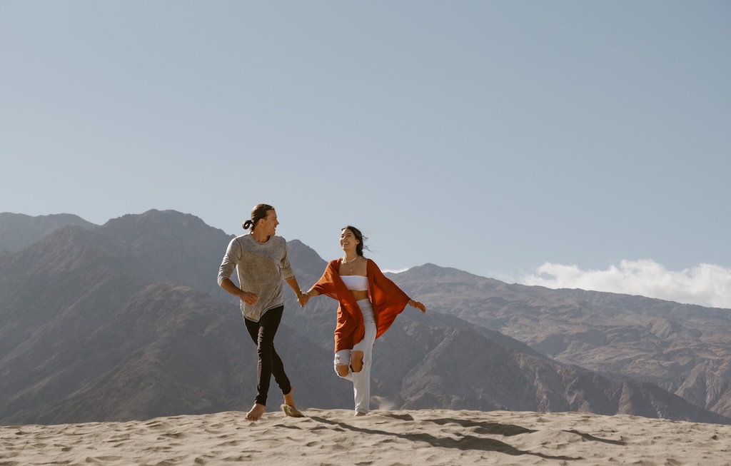A couple holding hands and running through the Mesquite Flats Sand Dunes in Death Valley, with mountains rising in the background.