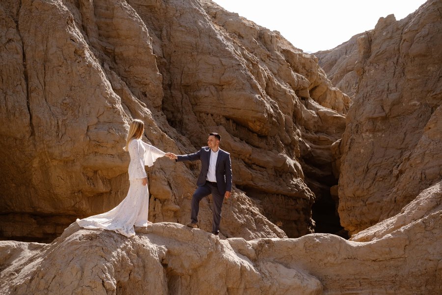 A couple standing on rocky terrain in a desert canyon, holding hands as they share a moment of connection against a dramatic natural backdrop.