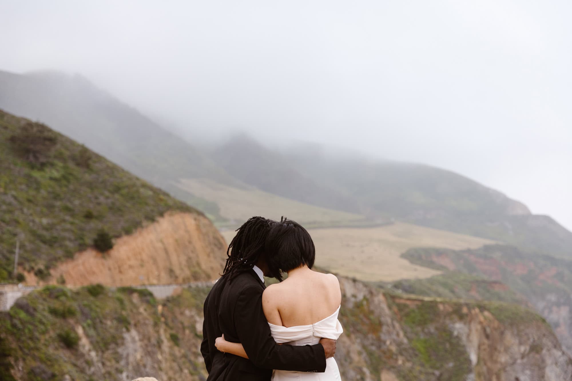 A couple embraces on a misty cliffside, overlooking rolling hills and winding roads in Big Sur.