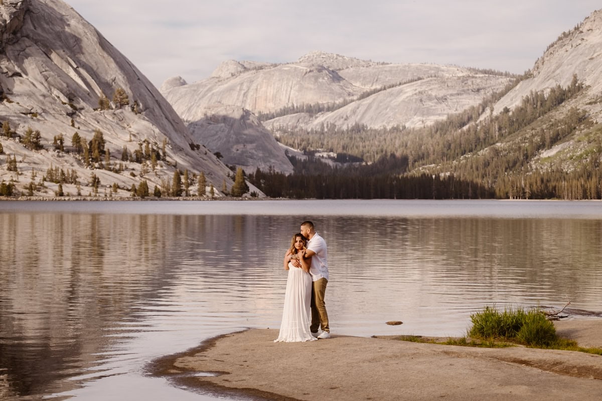 A couple stands together on a sandy edge of Tenaya Lake, with trees and mountains in Yosemite in the background.