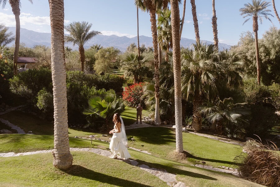 A bride walks gracefully through a lush garden surrounded by palm trees at The Oasis at Death Valley, with vibrant greenery and desert mountains in the distance under a bright blue sky.