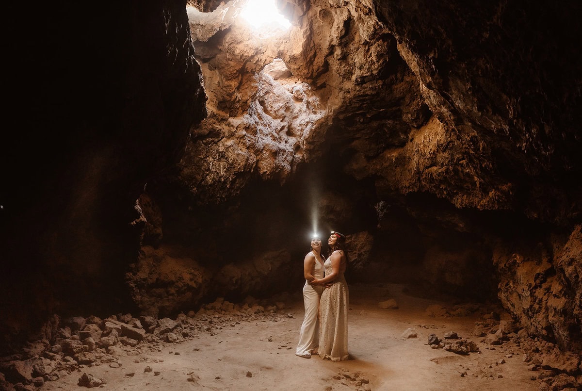 A couple eloping in the Mojave Desert stands inside a lava tube, with sunlight streaming through a natural opening above them.