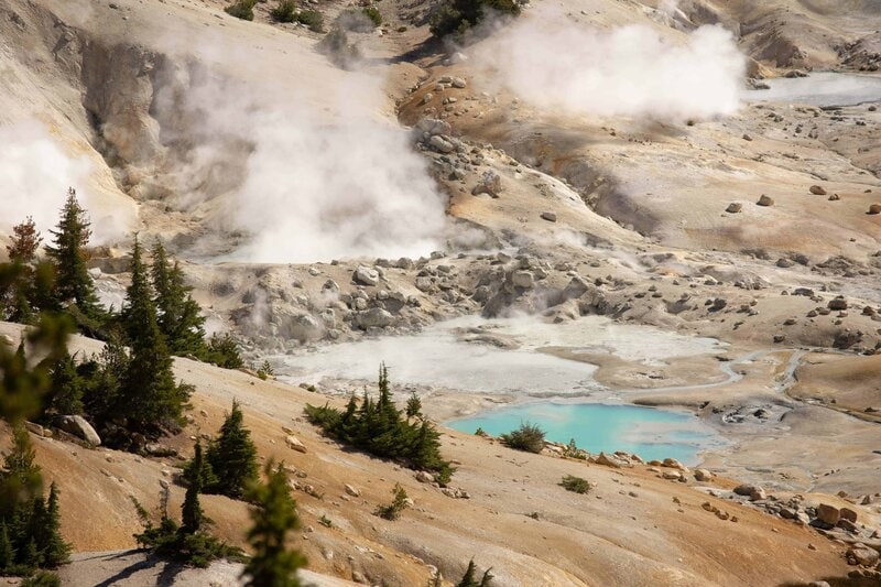 Steaming fumaroles rise from a rugged geothermal area with a turquoise hot spring, surrounded by barren slopes and scattered evergreen trees.